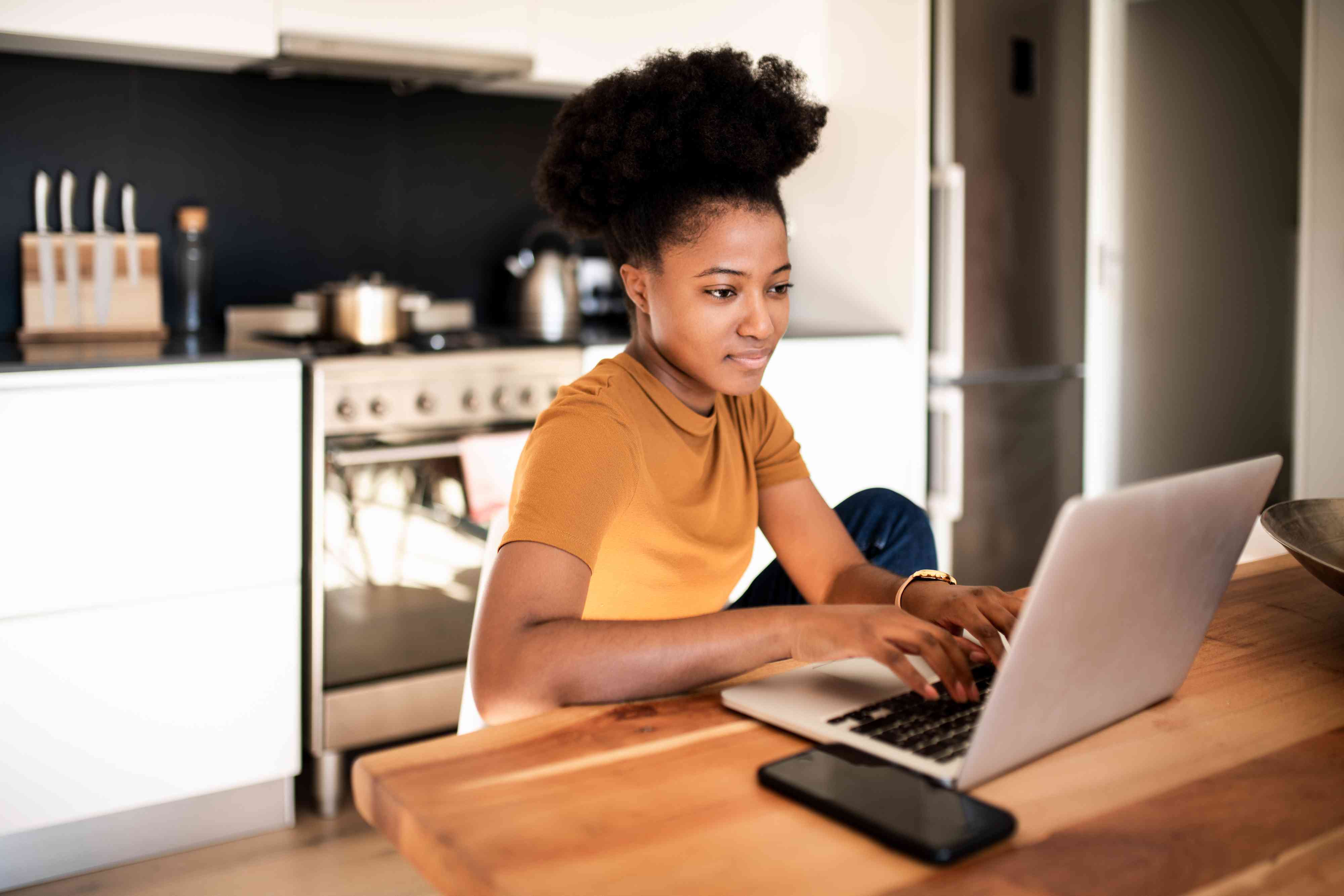 A woman sits at a kitchen table working on a laptop.